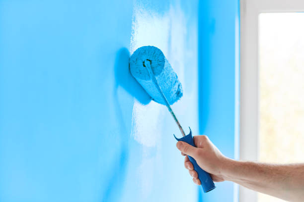 Человек красит стену в синий цвет малярным валиком.