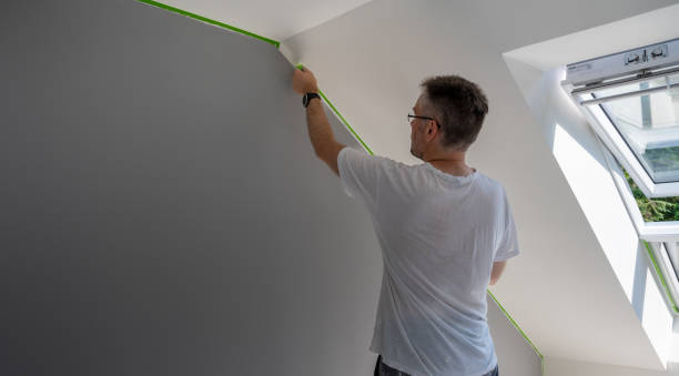 Мужчина приклеивает зеленую малярную ленту к стене, готовясь к покраске в помещении.